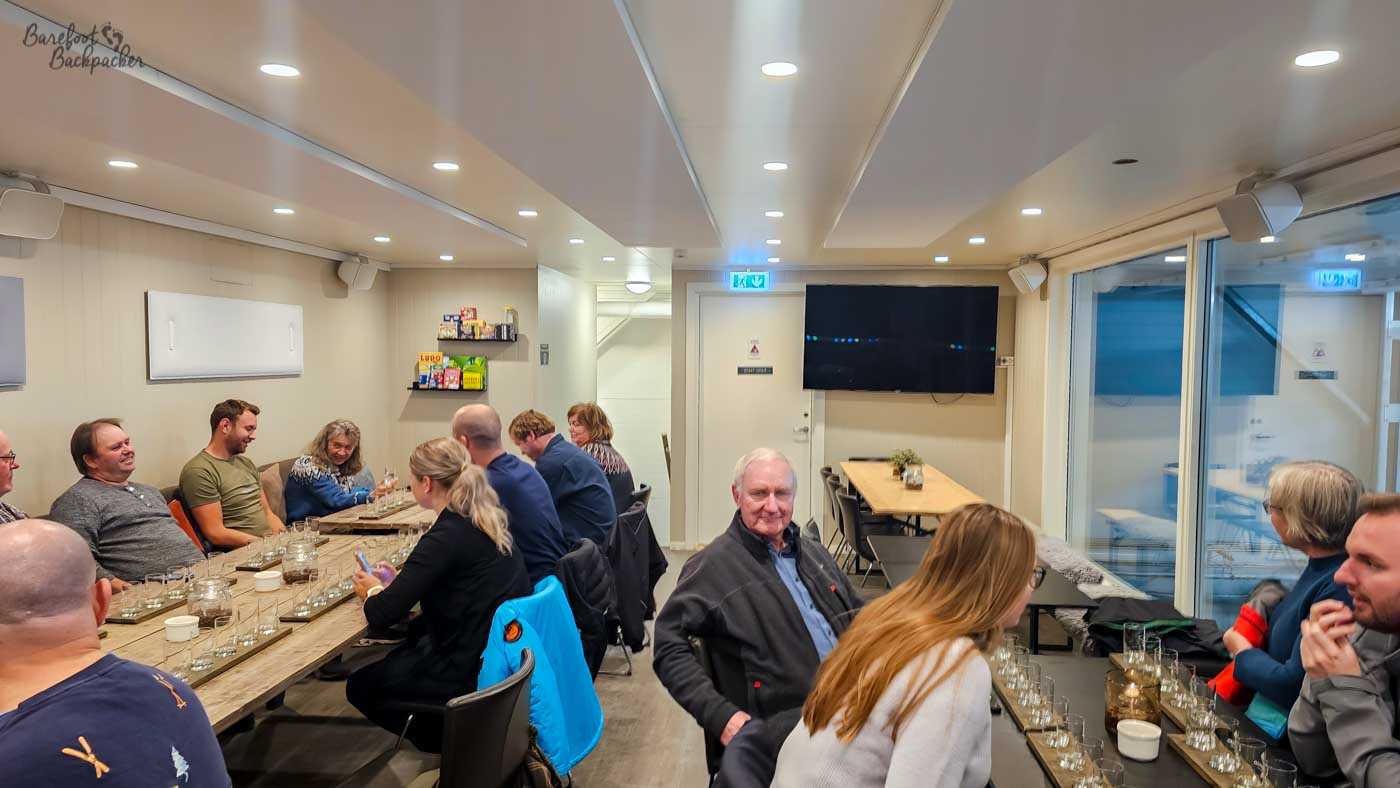 Inside a bright, modern tasting room, a group of people sit around long wooden tables, chatting and sampling drinks from rows of small glasses arranged in front of them. The room is warmly lit with recessed ceiling lights, contrasting with the cold environment outside, and shelves with snacks and a large wall-mounted screen add to the casual, social atmosphere. People of different ages are engaged in conversation, some smiling and leaning toward each other. The scene feels relaxed and convivial, a welcome indoor pause after time spent in the harsh Arctic landscape