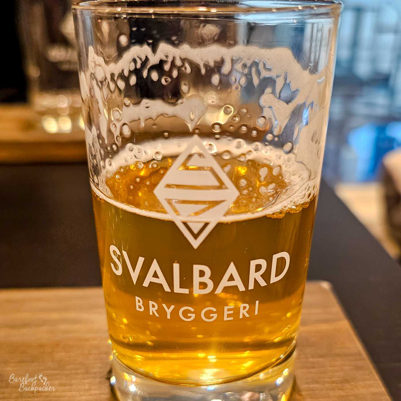 A close-up of a partially filled glass of golden beer shows bubbles clinging to the sides and a thin ring of foam near the top. The glass is branded with the text “SVALBARD BRYGGERI” and a geometric logo, clearly visible through the amber liquid. Warm indoor lighting gives the beer a rich, glowing colour, while the background is softly blurred, hinting at a bar or tasting setting. The image captures a simple, satisfying moment of refreshment after a day in the cold.