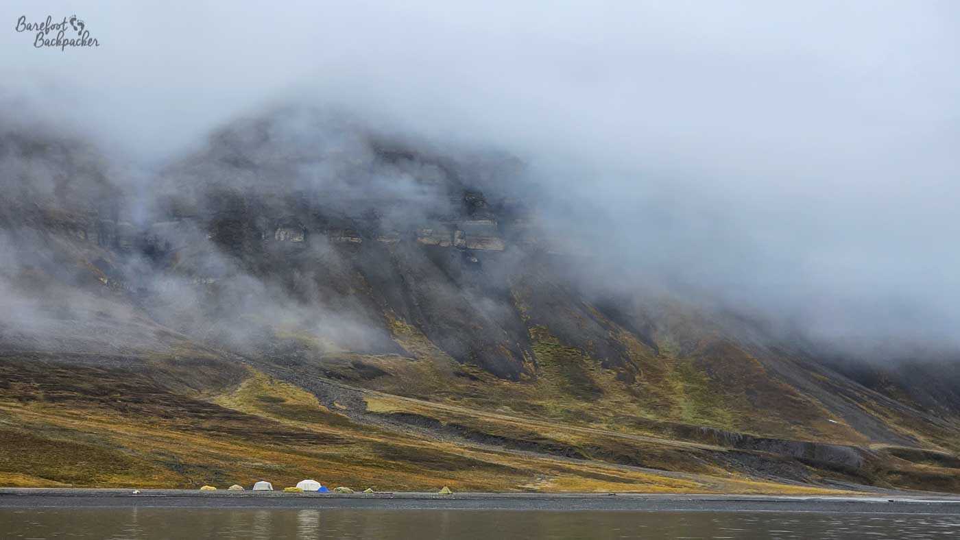 Low cloud clings to a steep mountainside, partially veiling dark rock and streaks of green vegetation beneath a shifting layer of mist. At the base of the slope, a narrow strip of shoreline hosts a small cluster of colourful tents pitched beside calm water, their bright hues standing out against the muted Arctic palette. The mountain looms above, its upper reaches disappearing into fog, giving the scene a quiet, slightly mysterious atmosphere. The still water in the foreground reflects the grey sky, adding to the sense of isolation and calm.