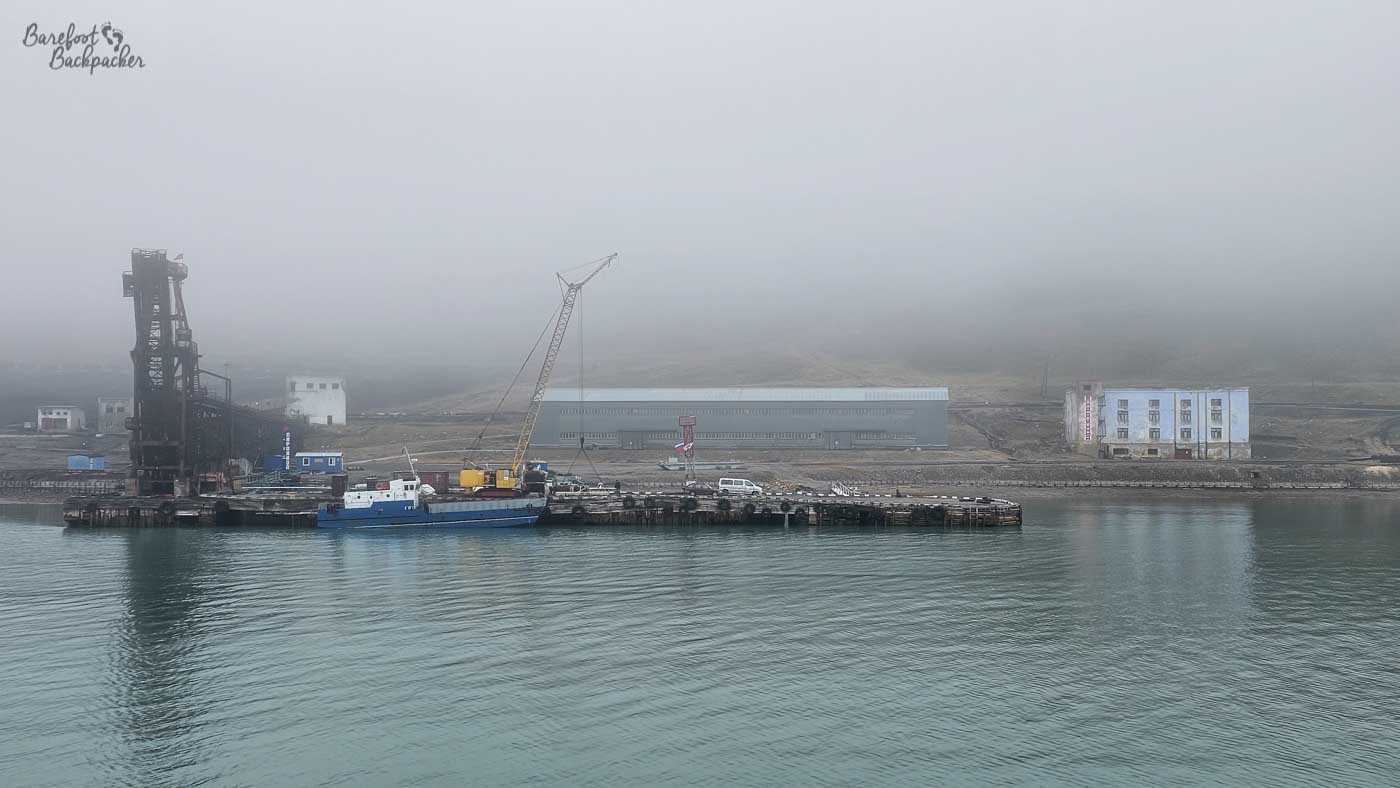 A mist-shrouded harbour scene with an old industrial loading structure and crane standing on a wooden pier. A small blue boat is moored alongside, while low buildings fade into the fog behind, giving the waterfront a quiet, almost ghostly atmosphere.