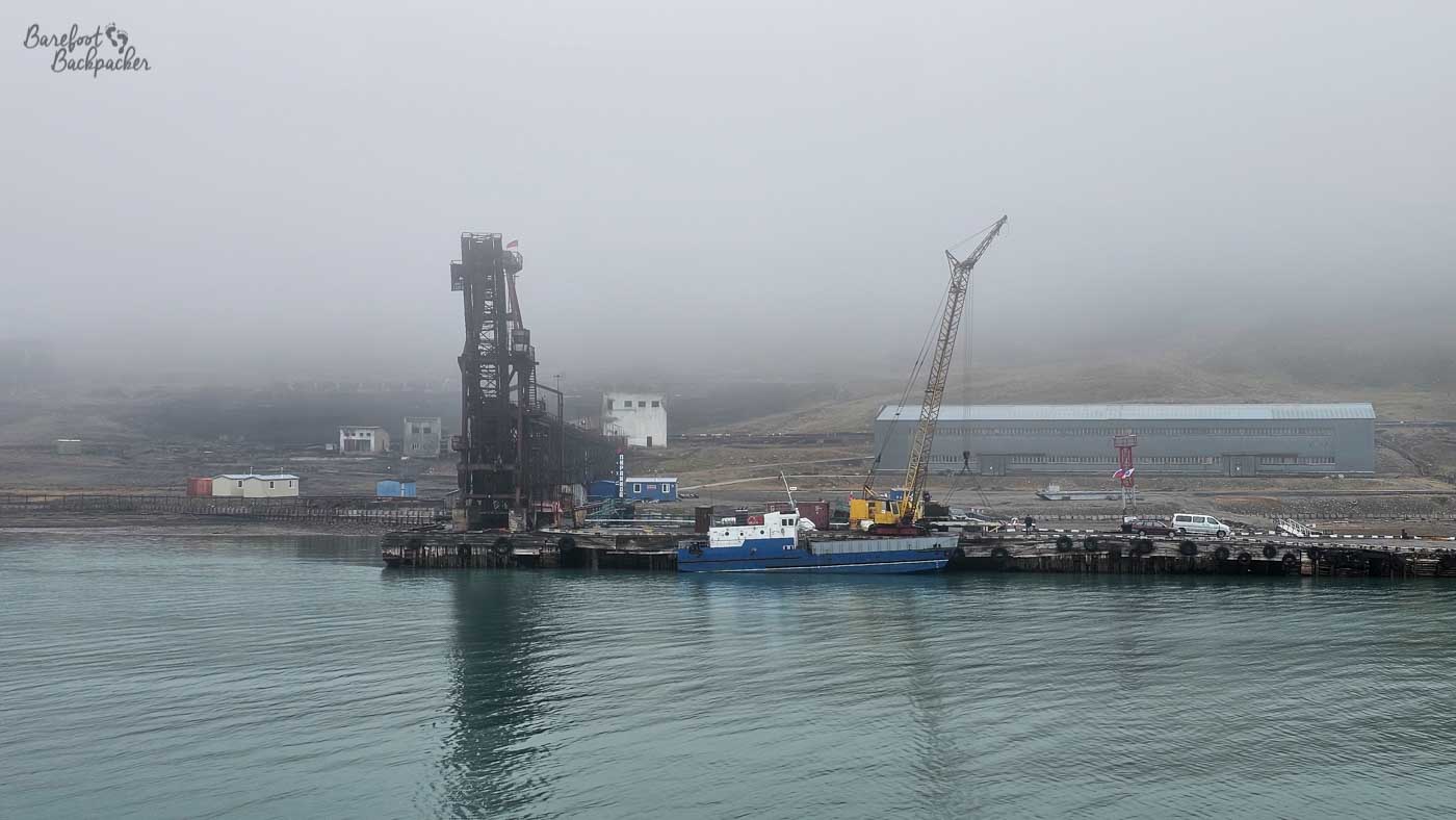 An industrial pier juts into pale, glassy water, dominated by a tall, dark metal loading structure and a crane rising beside it. A small blue-and-white vessel is moored alongside, dwarfed by the heavy machinery and the long warehouse building stretching behind. The entire scene is softened by thick fog, which obscures the background hills and gives the structures a faded, almost ghostly presence. The muted colours and quiet water create a stark, utilitarian Arctic harbour scene.