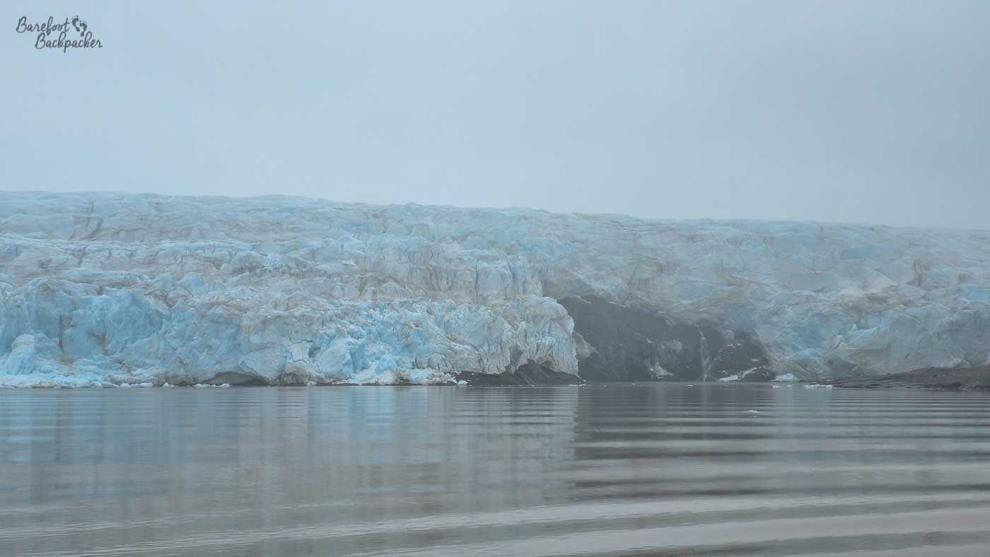 A wide glacier face spans the horizon, its blue-white ice broken into jagged ridges and crevasses that meet the calm sea. The water in the foreground is smooth, marked by gentle ripples that reflect the pale tones of the ice and sky. Mist hangs low over the glacier, blurring its upper edge and blending it into the overcast sky. The scene feels vast and silent, with the cold, textured ice dominating the landscape.