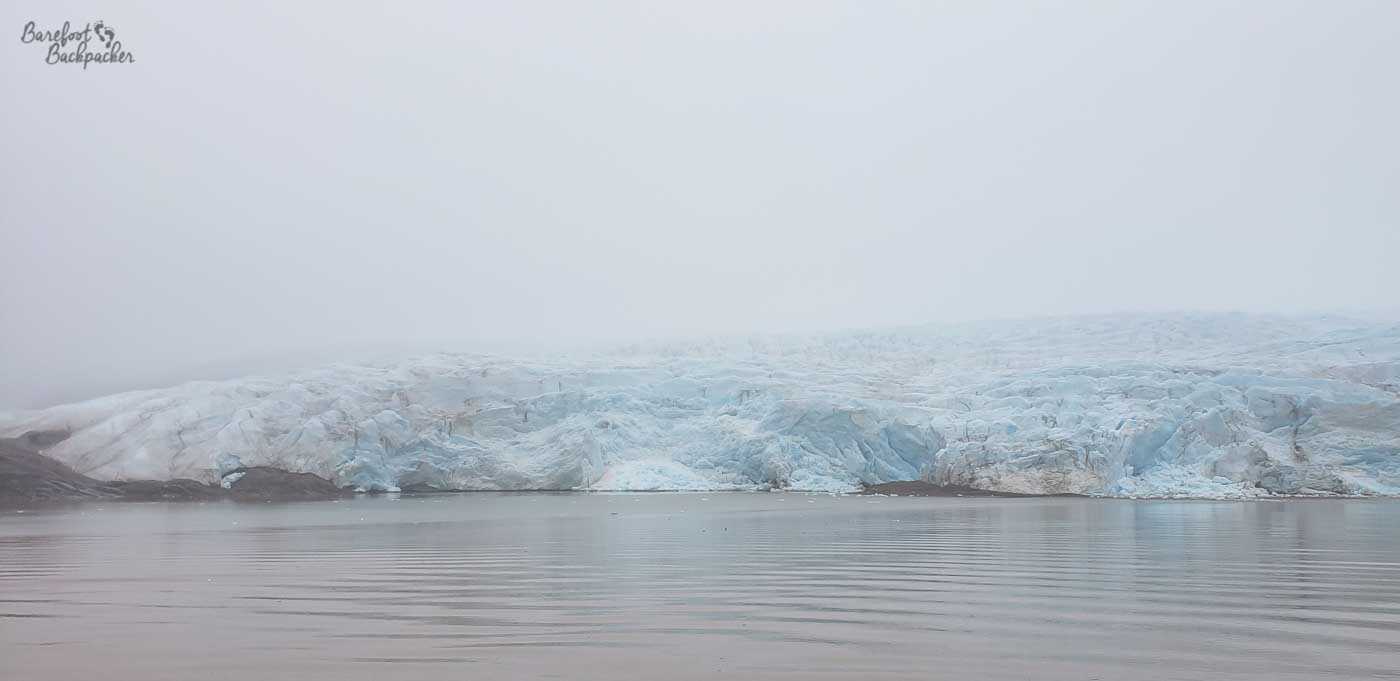 A wide, pale-blue glacier stretches across the horizon, its jagged ice front meeting calm, rippling water. Low cloud and fog soften the scene, muting the colours and blending ice, sky, and sea into a cool, monochrome landscape.