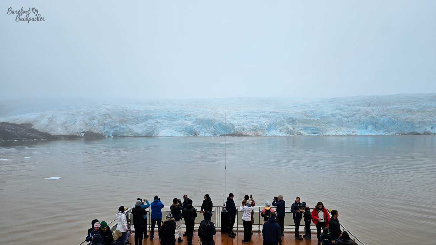 Passengers gather along the open deck of a ship, many bundled in jackets and hats as they face outward toward a distant glacier stretching across the water. Some people hold up phones or cameras, capturing the view, while others simply stand and watch. The glacier forms a long, pale backdrop under a grey, foggy sky, with the calm sea separating it from the ship. The human activity in the foreground contrasts with the immense, still landscape beyond.