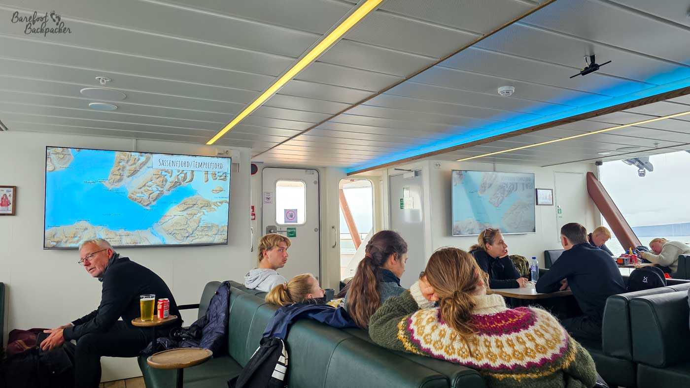 Inside a modern ferry lounge, passengers sit on cushioned benches and chairs, chatting, resting, or looking toward large wall-mounted screens displaying a map of the surrounding fjords. Warm lighting and clean, light-coloured interiors create a comfortable atmosphere, a contrast to the cold, foggy conditions outside. Drinks and small tables are scattered around, with people dressed in casual outdoor clothing. The scene captures a relaxed, communal moment during a journey through remote Arctic waters.