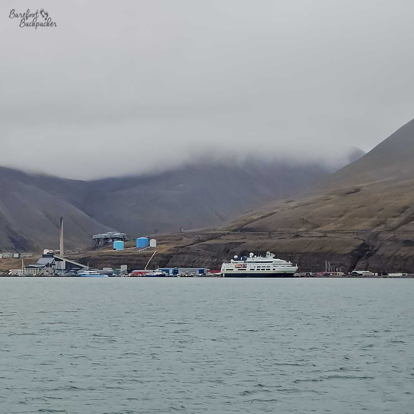 View of Longyearbyen from the sea, with clustered buildings lining the shoreline beneath steep, dark mountains. A large white ship is docked at the port, and low clouds hang over the peaks, reflecting softly on the calm Arctic water in the foreground.