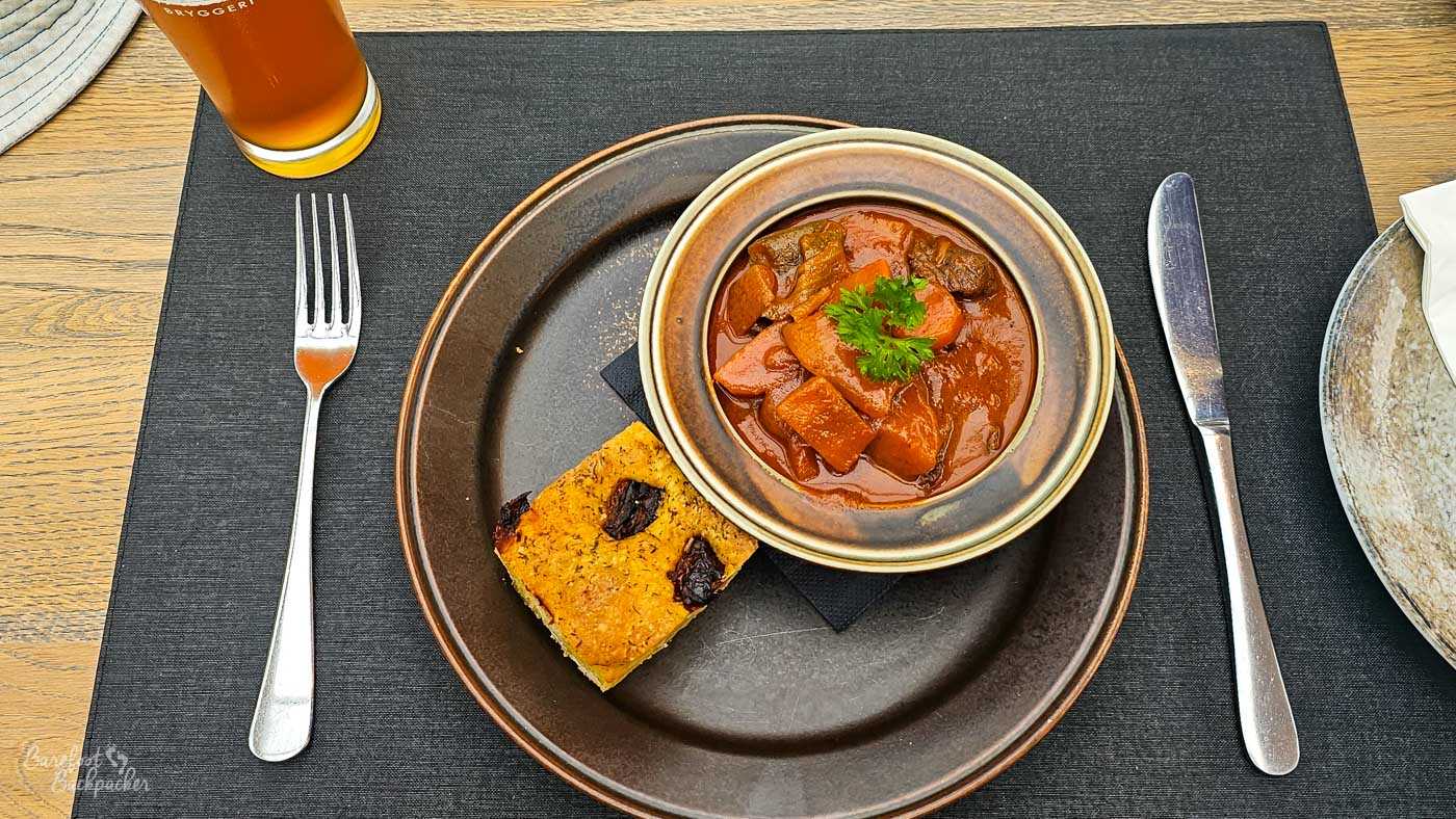 Top-down view of a rustic meal: a bowl of rich, reddish-brown stew filled with chunks of meat and vegetables, garnished with parsley, served on a dark plate alongside a slice of bread. A glass of amber beer sits just above the plate on a wooden table.