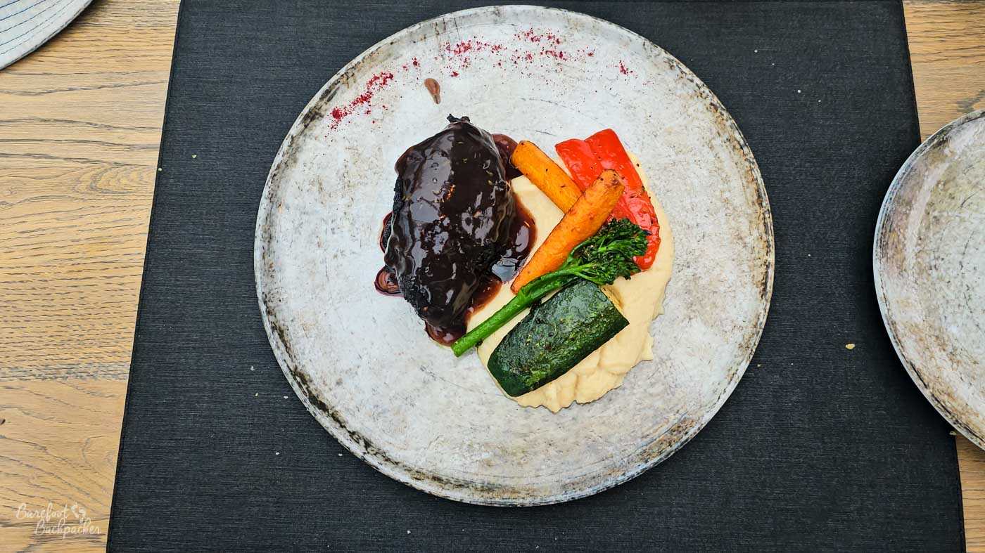 A neatly presented dish on a large ceramic plate: a piece of braised meat coated in glossy dark sauce, served with creamy mashed potatoes and a selection of vegetables including carrots, broccolini, red pepper, and courgette.