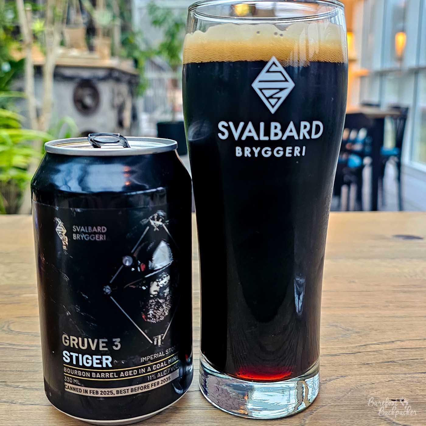 A tall glass of near-black stout with a thick tan head sits on a wooden table beside a matching Svalbard Bryggeri can labelled “Gruve 3 Stiger,” an imperial stout aged in a coal mine. The rich, opaque beer catches a hint of light at the base, while a softly blurred bar setting forms the background.