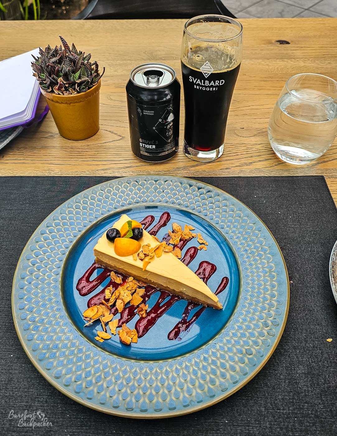 A neatly plated slice of cheesecake topped with berries and nuts sits on a patterned blue plate with berry sauce. Behind it are a glass and can of Svalbard Bryggeri stout, a glass of water, and a small potted succulent on the table.