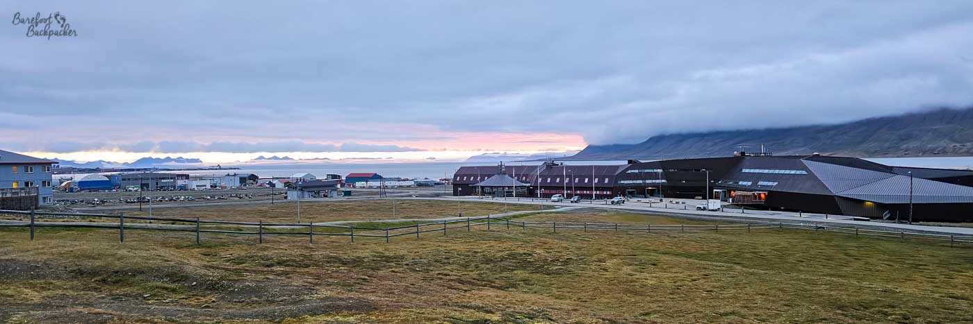 Wide view across Longyearbyen’s outskirts toward the sea at sunset, with low buildings and roads stretching across a grassy foreground. The sky glows pink and orange beneath heavy clouds, with distant mountains silhouetted along the horizon.