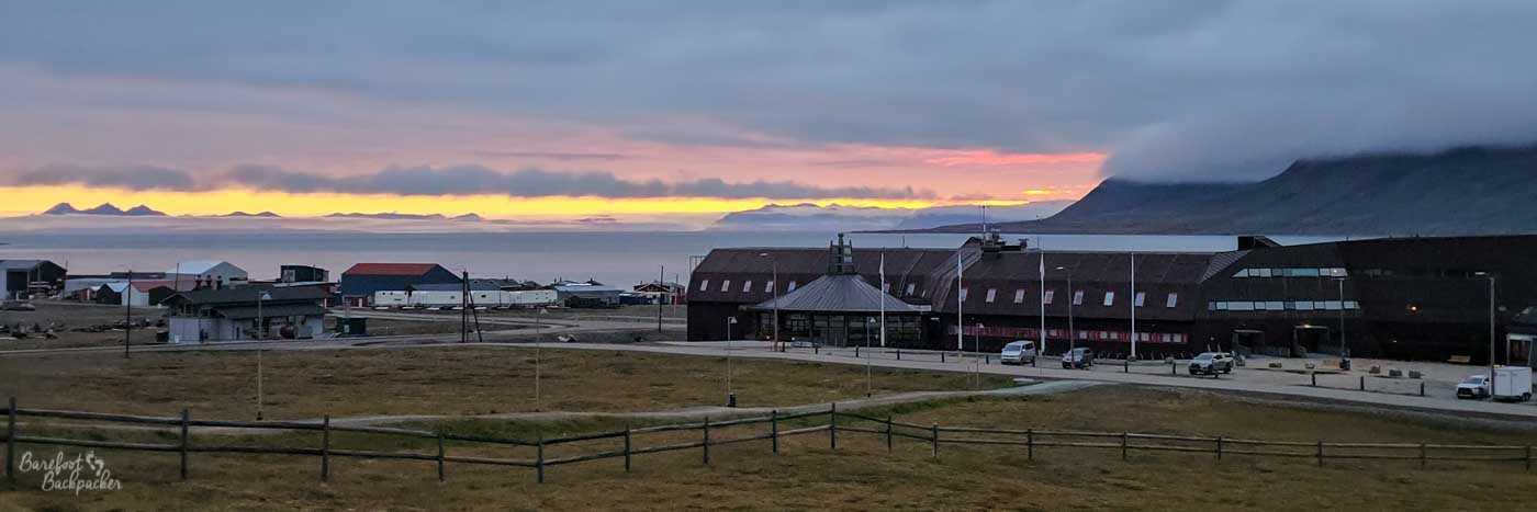 A wide view over Longyearbyen at sunset, with low buildings and a large dark-roofed structure in the foreground. A thin band of glowing orange light sits beneath heavy clouds above the calm fjord and distant mountain silhouettes.