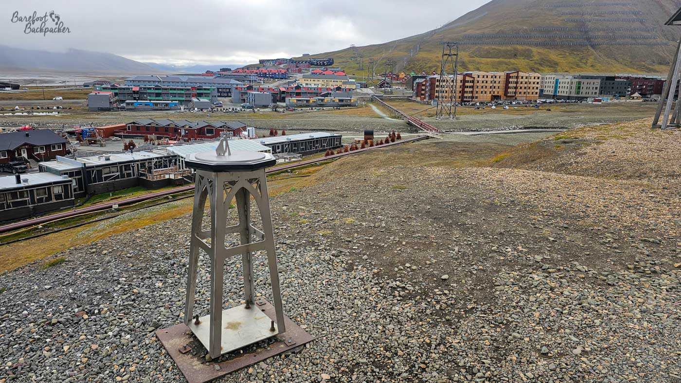 A small metal structure stands on a rocky hillside overlooking Longyearbyen, with old industrial pipelines and infrastructure cutting across the landscape below, and rows of buildings backed by steep, sparsely vegetated slopes.