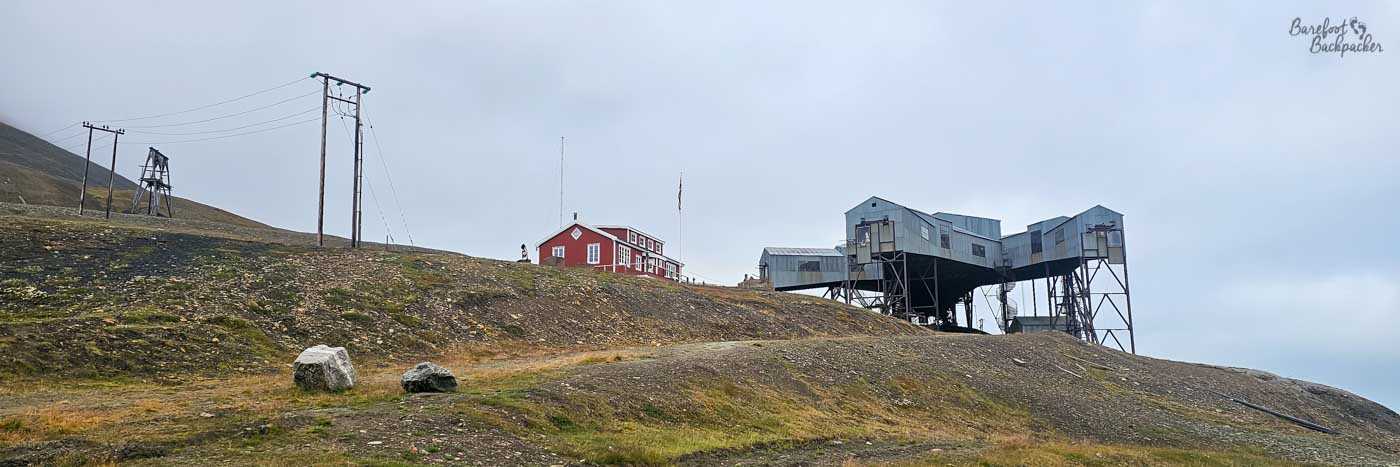 A sparse Arctic hillside stretches across the frame, dotted with patches of low vegetation and scattered rocks beneath a pale, overcast sky. Perched along the ridge are a handful of isolated buildings: a striking red wooden house stands out vividly against the muted landscape, while nearby, a cluster of industrial-looking structures on stilts—metallic and angular—suggest mining or research activity. Wooden utility poles march across the slope, their wires cutting thin lines through the empty air. The scene feels remote and functional, a place where human presence is small but purposeful against the vast, exposed terrain.