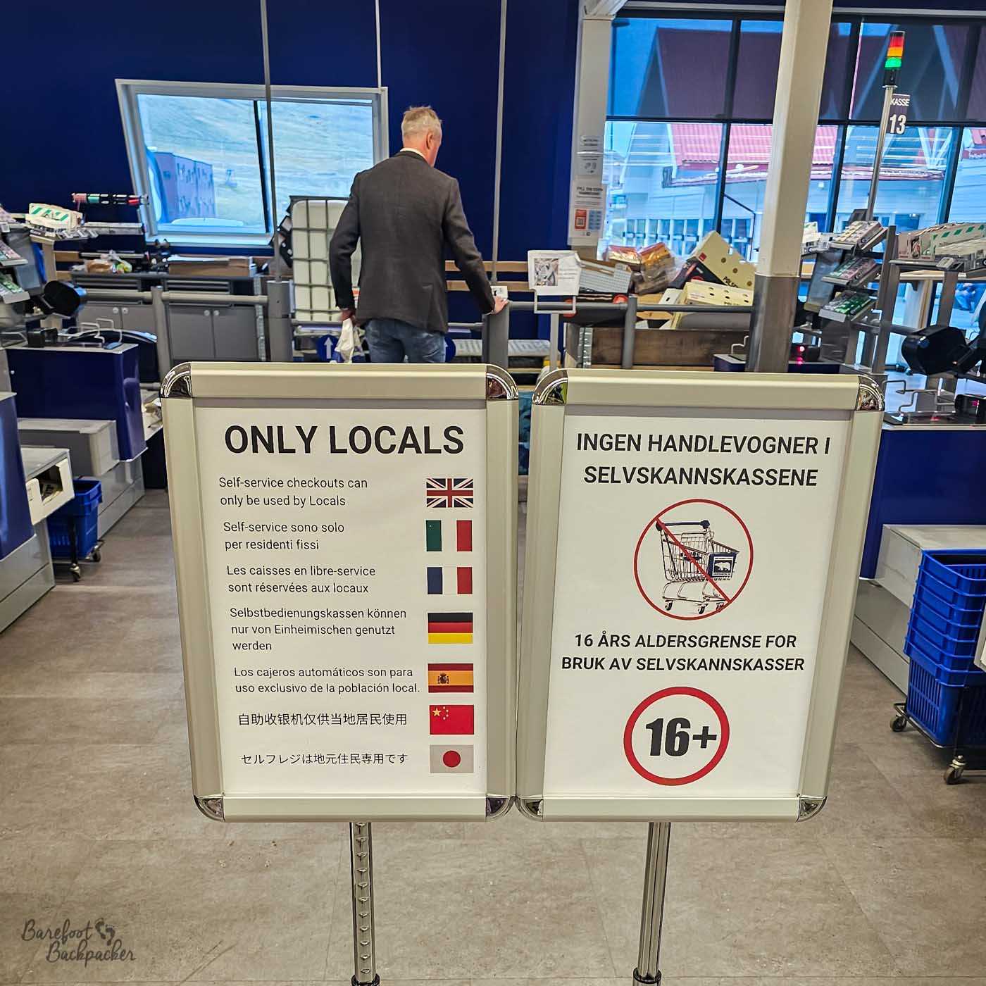 Signs at a supermarket self-checkout area state that the machines are for local residents only, with multilingual text and flags. A second sign indicates a 16+ age limit and no shopping trolleys, while a customer stands beyond the barrier.