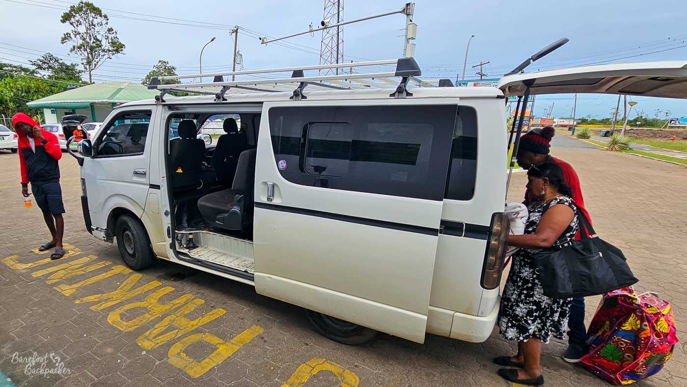 A white minibus with its side door open is parked at a roadside stop, as passengers load bags and prepare to board. A woman in a patterned dress lifts a large, colourful bag into the back while another person stands nearby, and a man in a red hoodie lingers off to the side holding a drink. The pavement is marked with faded yellow lettering, and low buildings, utility poles, and a tangle of wires frame the background. The moment feels busy yet routine—a shared departure point in everyday local travel.