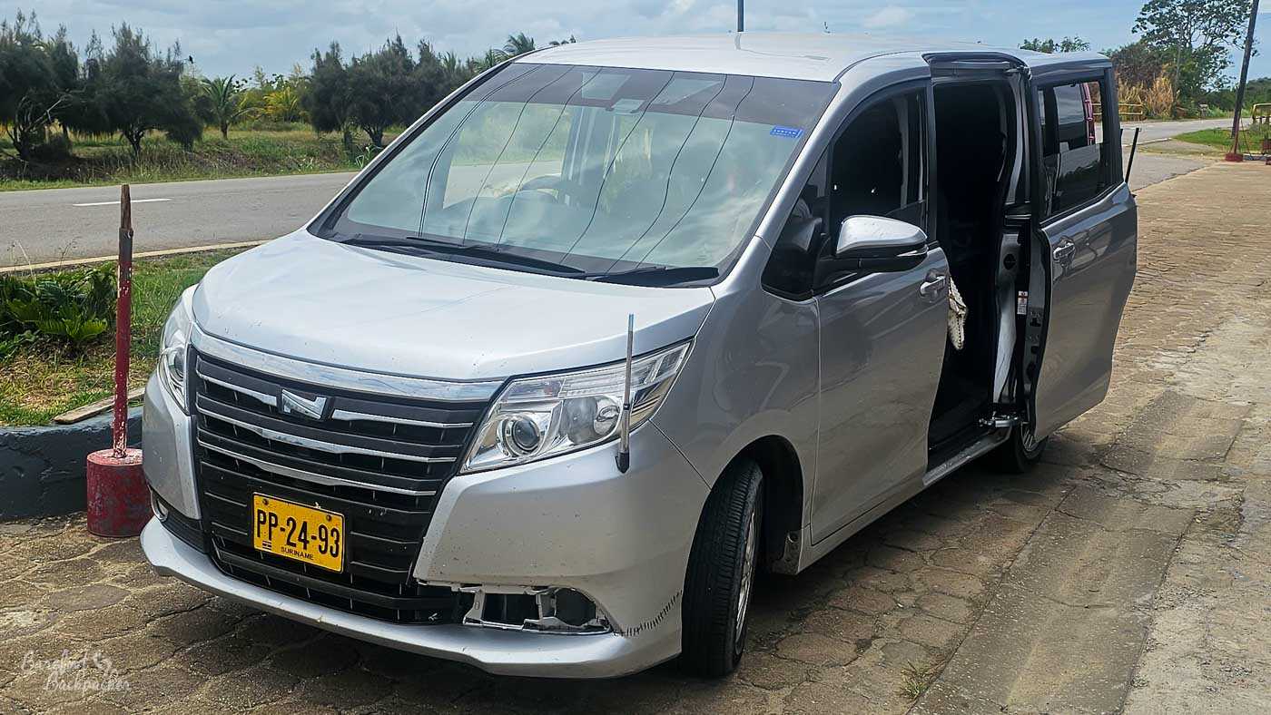 A silver minivan is parked on the side of a quiet roadside, its sliding door open to reveal the dark interior, ready for passengers or luggage. The vehicle shows signs of wear, with scuffs and a slightly damaged front bumper, hinting at long days on rough roads. A strip of grass and low vegetation separates the road from a line of small trees and palms in the background, under a hazy, overcast sky. The scene captures a pause in transit—practical, unglamorous, but essential to the journey.