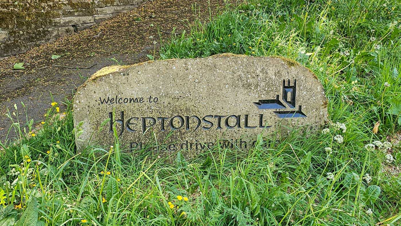 A carved stone welcome sign marking the entrance to Heptonstall village, partly surrounded by long grass and wildflowers.