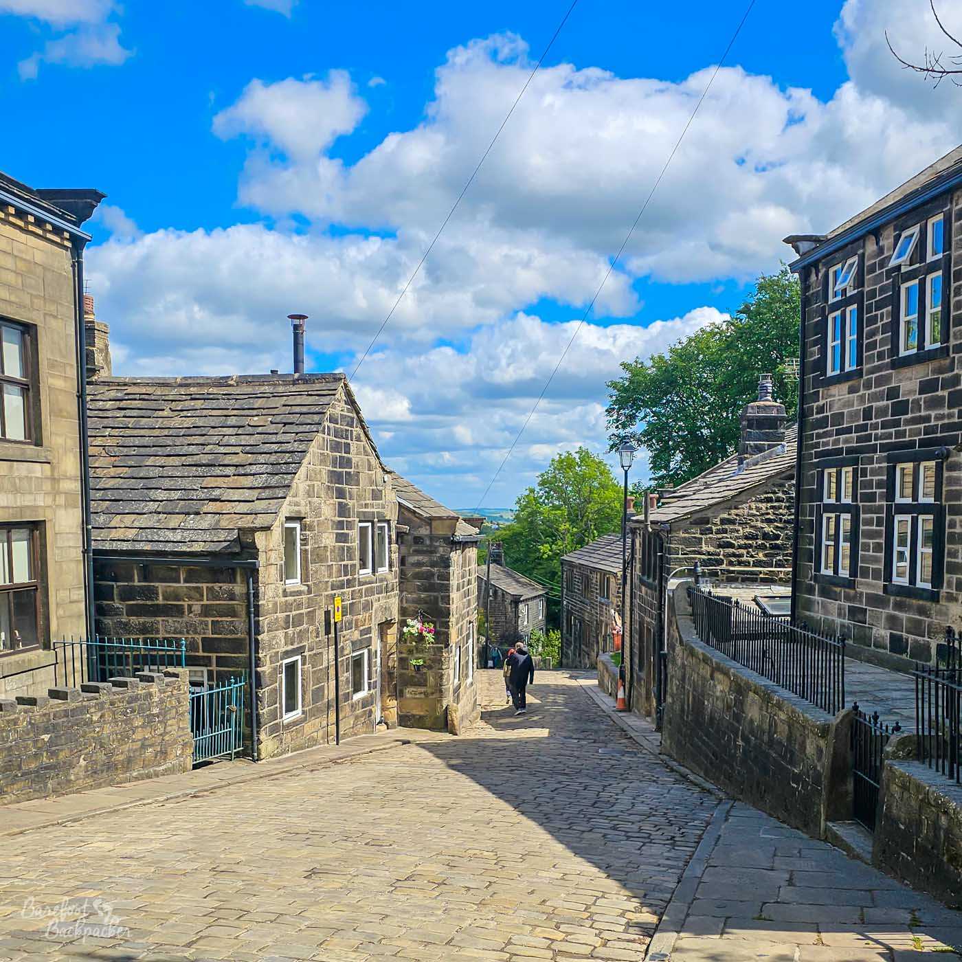 A steep cobbled street lined with traditional stone cottages and buildings in Heptonstall village, descending towards distant countryside beneath a bright blue sky filled with white clouds.