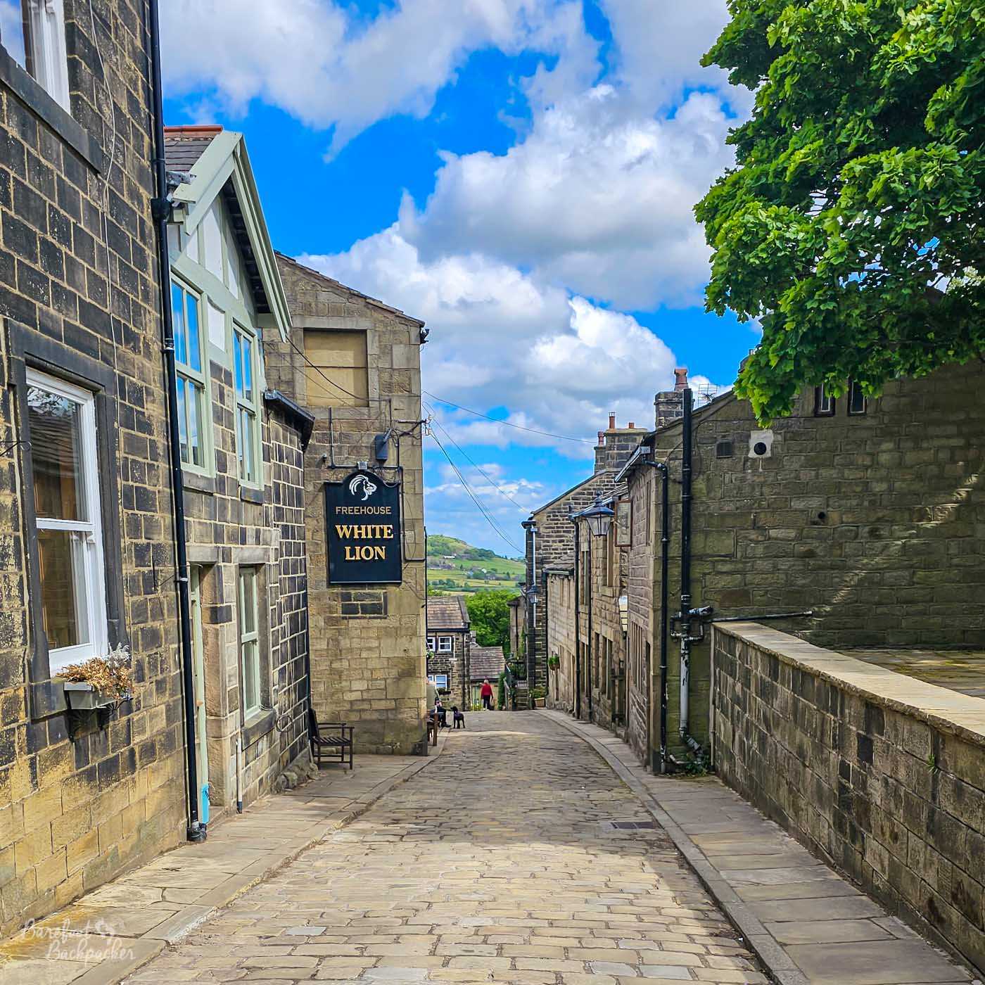A cobbled street lined with stone cottages and buildings in Heptonstall village, with the White Lion pub sign hanging above the road and countryside visible in the distance.