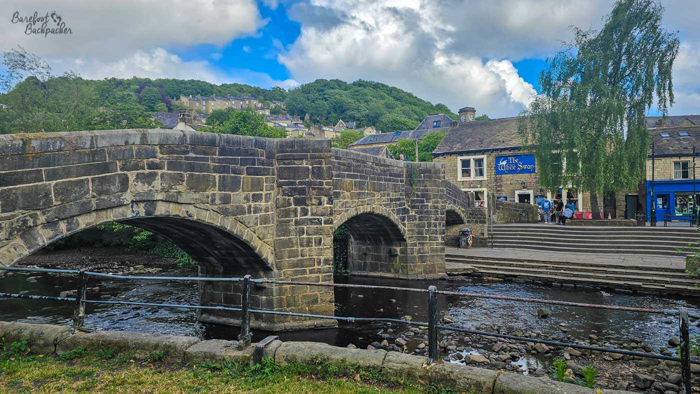 The historic stone bridge over the River Hebden spans three arches beside the White Swan pub, with trees and hillside houses rising behind the town centre.
