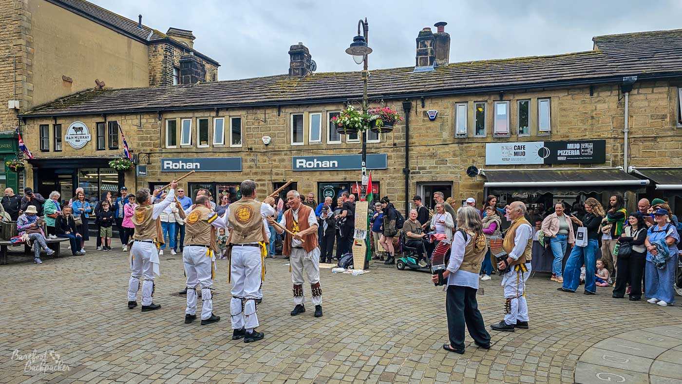 Morris dancers perform in Hebden Bridge's market square while shoppers and visitors gather in a circle to watch, with stone-fronted shops forming the backdrop.