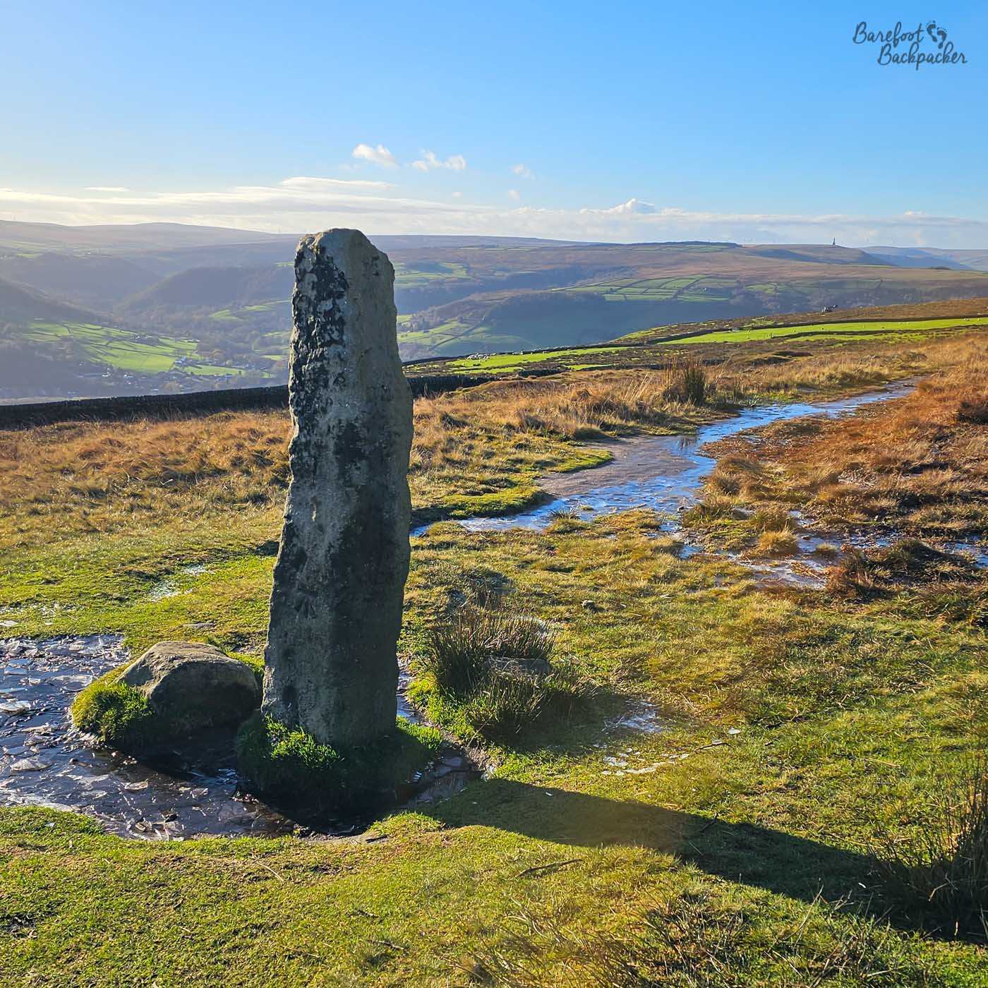 An ancient standing stone beside a muddy moorland path overlooks the Calder Valley, with Stoodley Pike visible on the distant skyline in warm late afternoon sunshine.