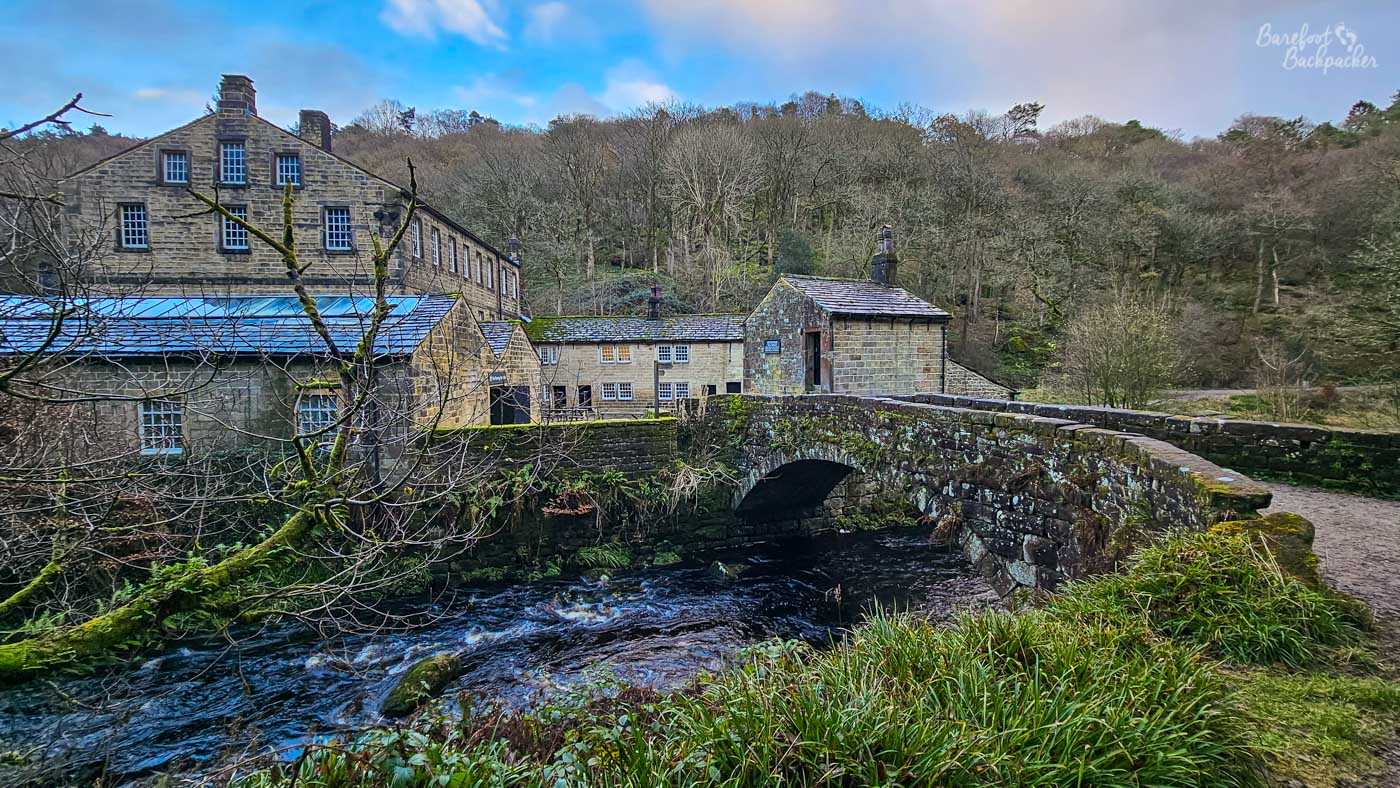 Gibson Mill beside the Hebden Beck, viewed across a stone bridge with woodland rising behind the historic mill buildings.