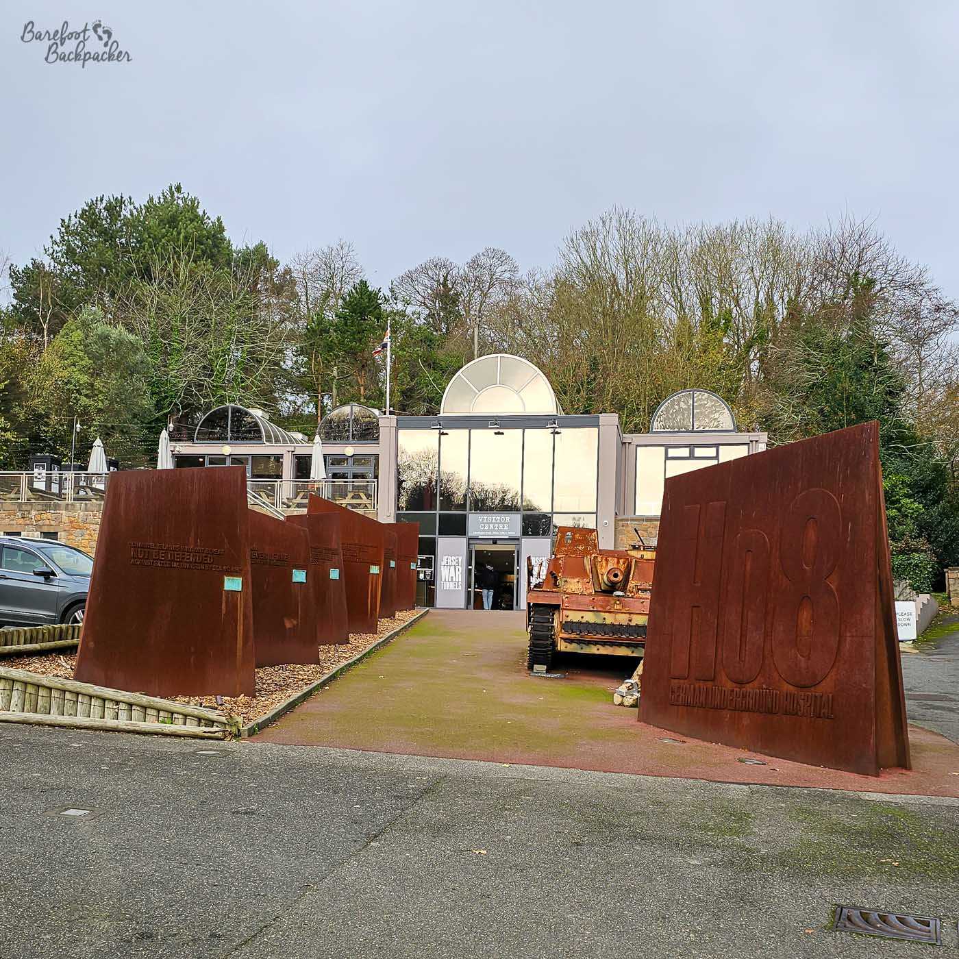 A view of the exterior of the Jersey War Tunnels with a large rusty metal sculpture in the foreground. The sculpture has the letters 'H08' and 'GERMAN UNDERGROUND HOSPITAL' cut out of it. To the left of the sculpture are several other similar, smaller rusty metal panels arranged in a line. Behind the sculptures is the entrance to the Jersey War Tunnels, a museum housed in a former German hospital. The building has a modern glass facade and a curved roof with skylights. A military tank is parked outside the entrance. A sign indicates 'VISITOR CENTRE' and 'JERSEY WAR TUNNELS'. The sky is overcast and the trees in the background are bare, suggesting it is autumn or winter. The overall impression is one of historical commemoration and a stark reminder of the past.