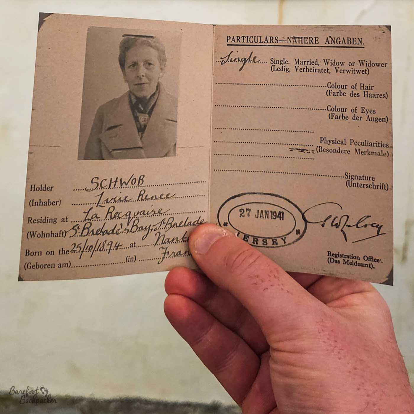 The image shows a vintage document, likely an identification or registration card from 1941. The document is partially held in a person's hand, obscuring some details. The card includes a black-and-white photograph of a woman in the upper left corner. Below the photograph, there are handwritten fields for details like the holder's name ('SCHWOB Lucie Renee'), address ('La Requaire'), and date and place of birth ('25/10/1894' at 'Nant, Fran'). On the right side of the card, under the heading 'PARTICULARS – NÄHERE ANGABEN', there are fields for marital status (indicated as 'Single'), hair and eye colour, and physical peculiarities, although these fields are mostly blank. The card is stamped with a seal that reads '27 JAN 1941 JERSEY.' A signature labelled 'Waley' and the text 'Registration Office. (Das Meldeamt).' are visible at the bottom right.