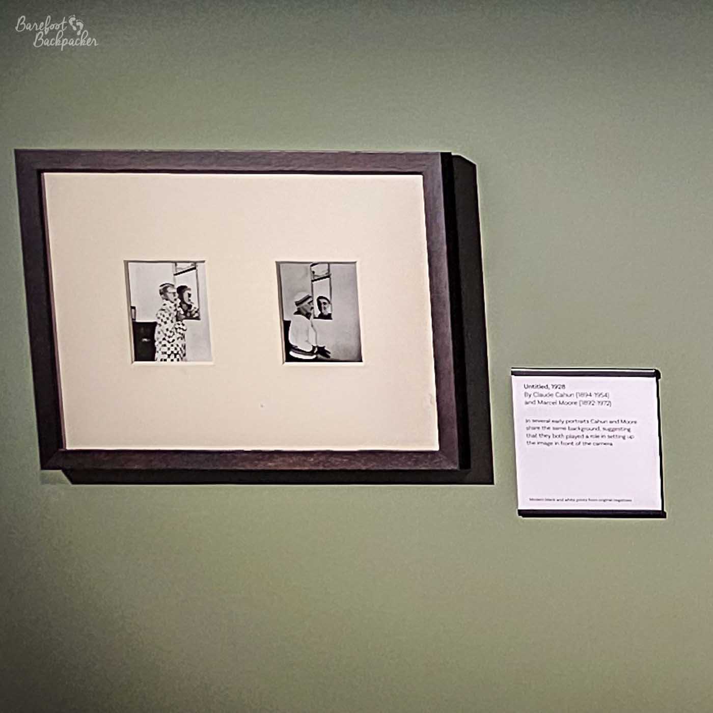 This image shows a framed artwork on a pale green wall. The artwork consists of a dark brown frame with a thick, light-coloured mat. Inside the mat, two small black and white photographs are displayed side-by-side. The photograph on the left shows a person in a patterned outfit looking out of frame. The photograph on the right shows a person in a sailor-like uniform looking towards the viewer. To the right of the artwork is a small white label with black text, providing information about the piece. The label reads 'Untitled, 1928 By Claude Cahun (1894-1954) and Marcel Moore (1892-1972)'. Below this, there is a descriptive text that mentions 'several early portraits' and how Cahun and Moore shared the same background, suggesting they both played a role in setting up the image in front of the camera. At the very bottom of the label, it states 'Modern black and white prints from original negatives'. The overall presentation suggests a museum or gallery setting, highlighting historical photographic works.