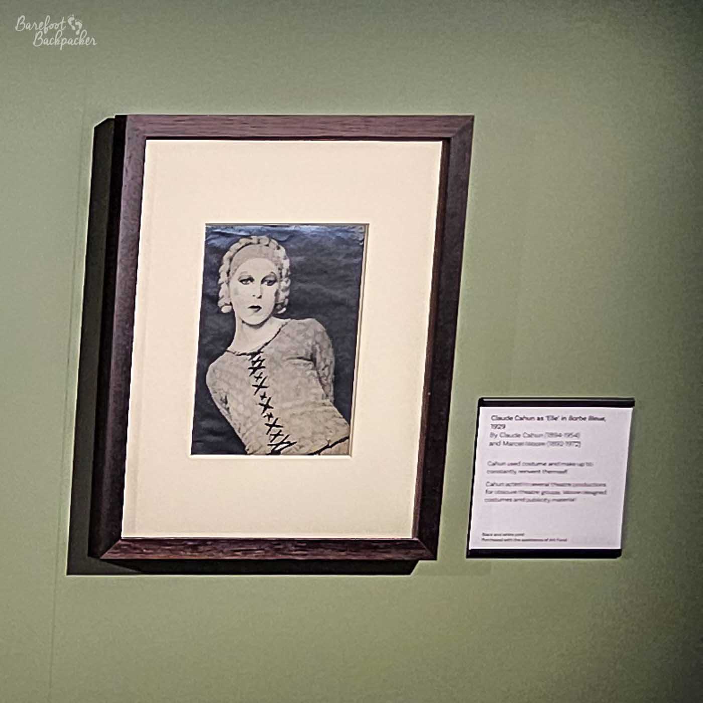 A framed photograph of Claude Cahun as 'Elle' in 'Sorbe Bleue' (1929) is displayed on a light green wall. The photograph, a black and white image, features Cahun with elaborate curly hair, a headband, and makeup. They are wearing a top with a criss-cross lacing pattern on the front. The photograph is matted with a thick cream-coloured border and framed in a dark brown wooden frame. To the right of the framed photograph, a small white label with black text provides information about the artwork. The label reads: Claude Cahun as 'Elle' in Sorbe Bleue, 1929 By Claude Cahun (1894-1954) and Marcel Moore (1892-1972). Cahun used costume and make-up to constantly convert themself. Cahun acted in several theatre productions for obscure theatre groups, labour designed costumes and publicity material. Black and white print. Purchased with the assistance of Art Fund.
