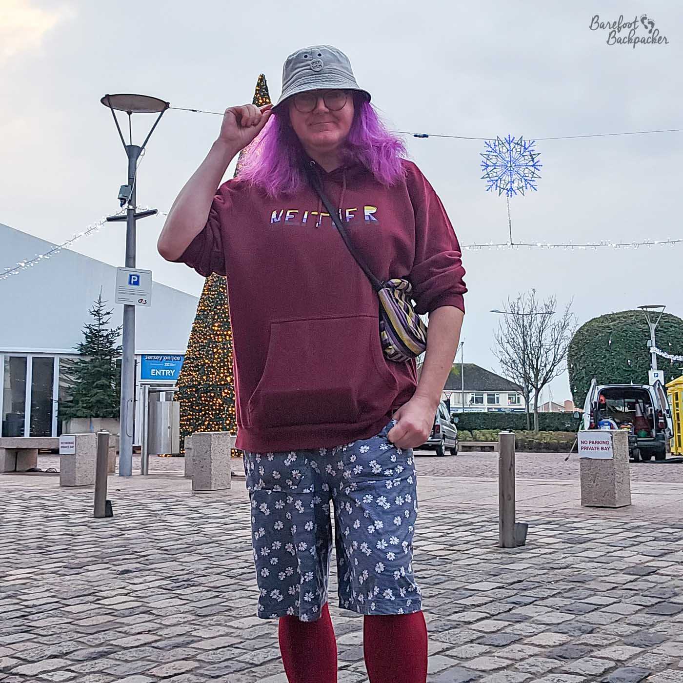 In a full-body shot, a person with purple hair poses in an outdoor setting. The person wears a grey bucket hat, glasses, a maroon hoodie with the word