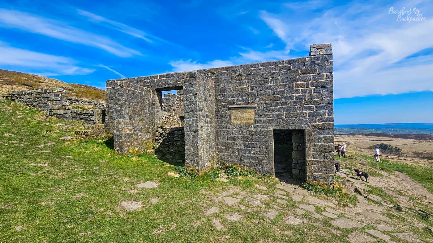 A side view of the ruined farmhouse at Top Withens shows its surviving stone walls overlooking the expansive Yorkshire moors, with walkers and a dog nearby.