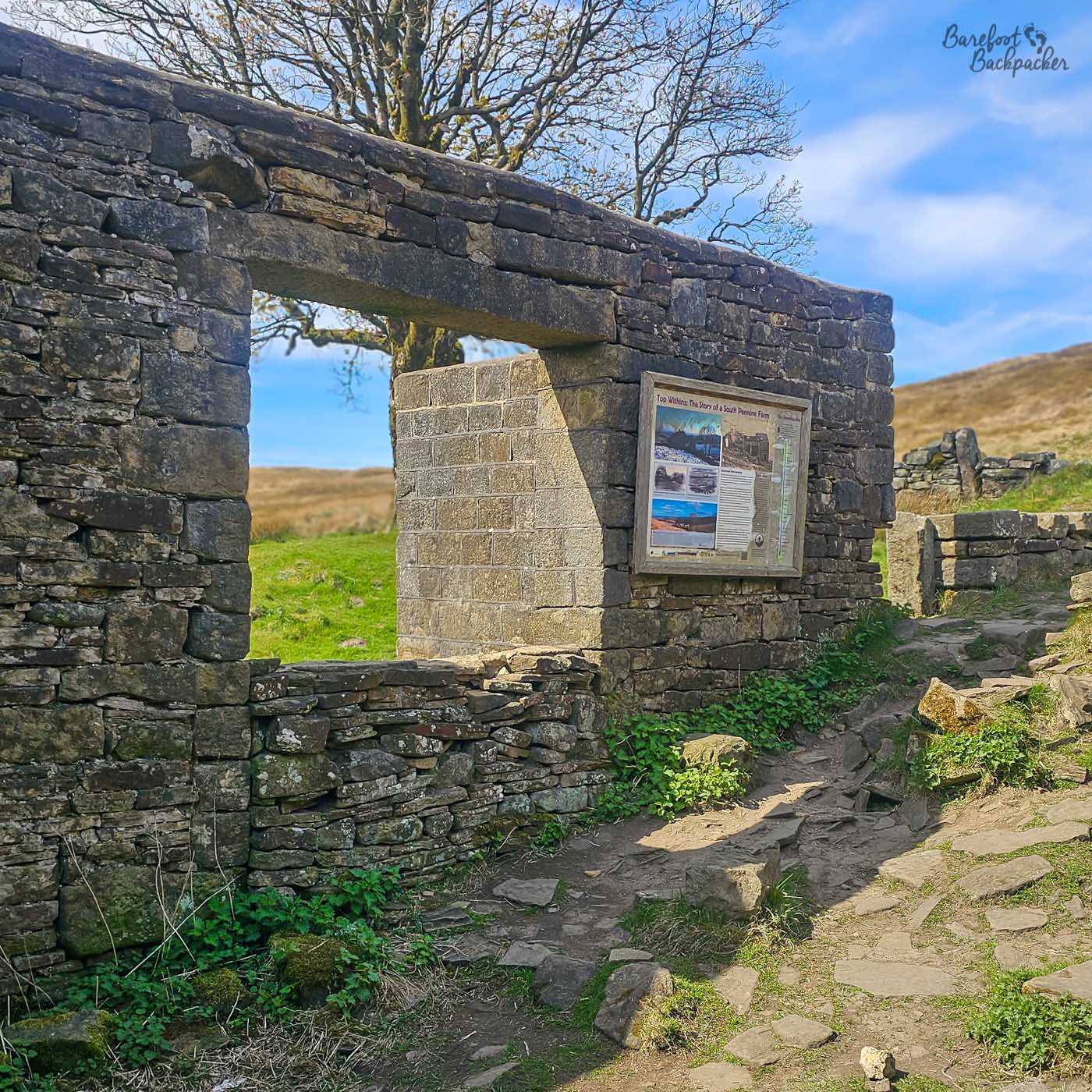 The stone entrance to the ruined farmhouse at Top Withens stands beside an information board, with rough moorland paths and dry-stone walls surrounding the isolated building.