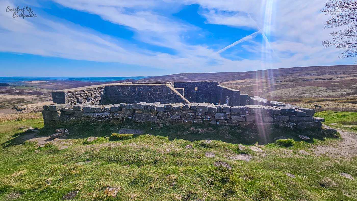 The ruined stone farmhouse known as Top Withens sits alone on the open Yorkshire moor, surrounded by rolling hills beneath a bright blue sky streaked with high cloud.
