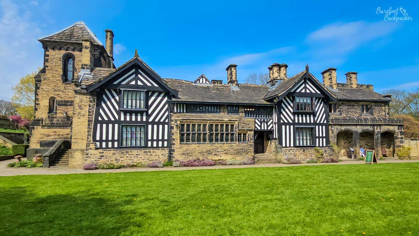 The distinctive black-and-white timbered façade of historic Shibden Hall stands behind a neatly kept lawn, with stone walls, gables and a square tower beneath a blue sky.