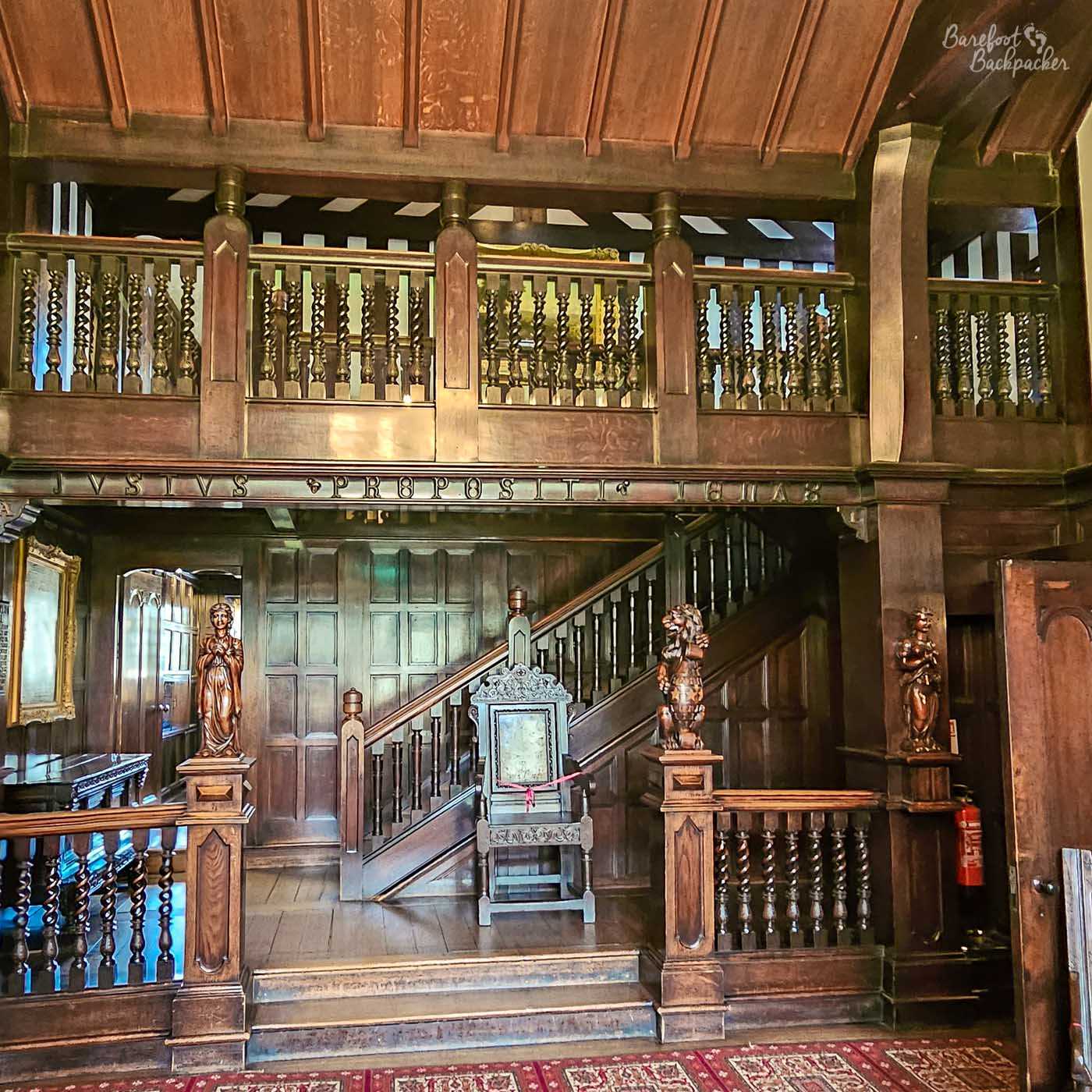 The richly panelled entrance hall of Shibden Hall features a carved wooden gallery, staircase, statues and an ornate ceremonial chair beneath a timber ceiling.