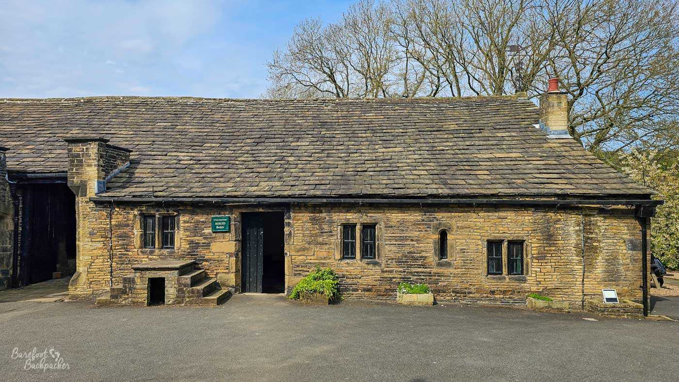 The long stone Aisled Barn at Shibden Hall has small mullioned windows, a central doorway and a traditional stone-slate roof.