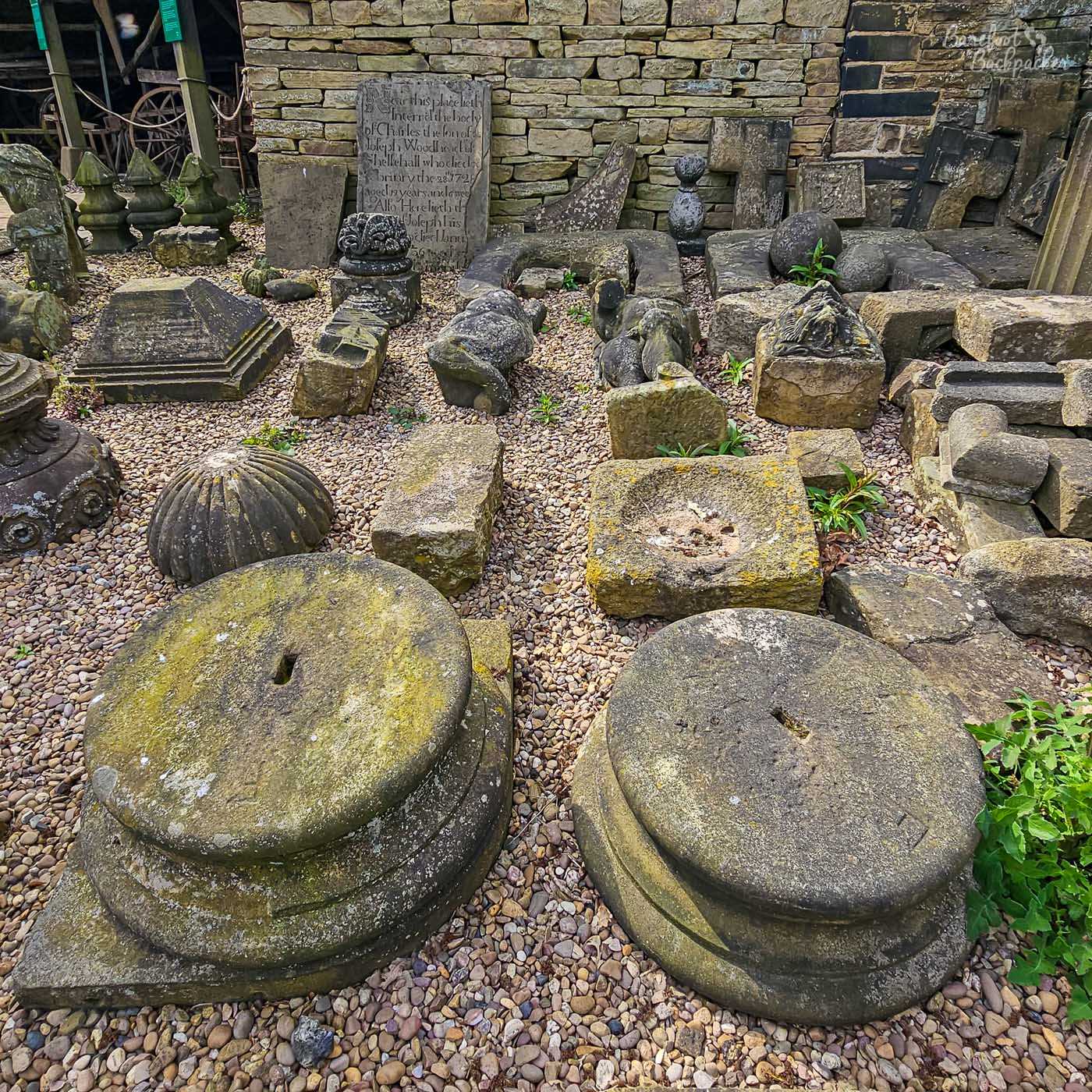 Historic carved stone fragments, finials and architectural details are displayed together in a gravel courtyard beside a stone wall at Shibden Hall.