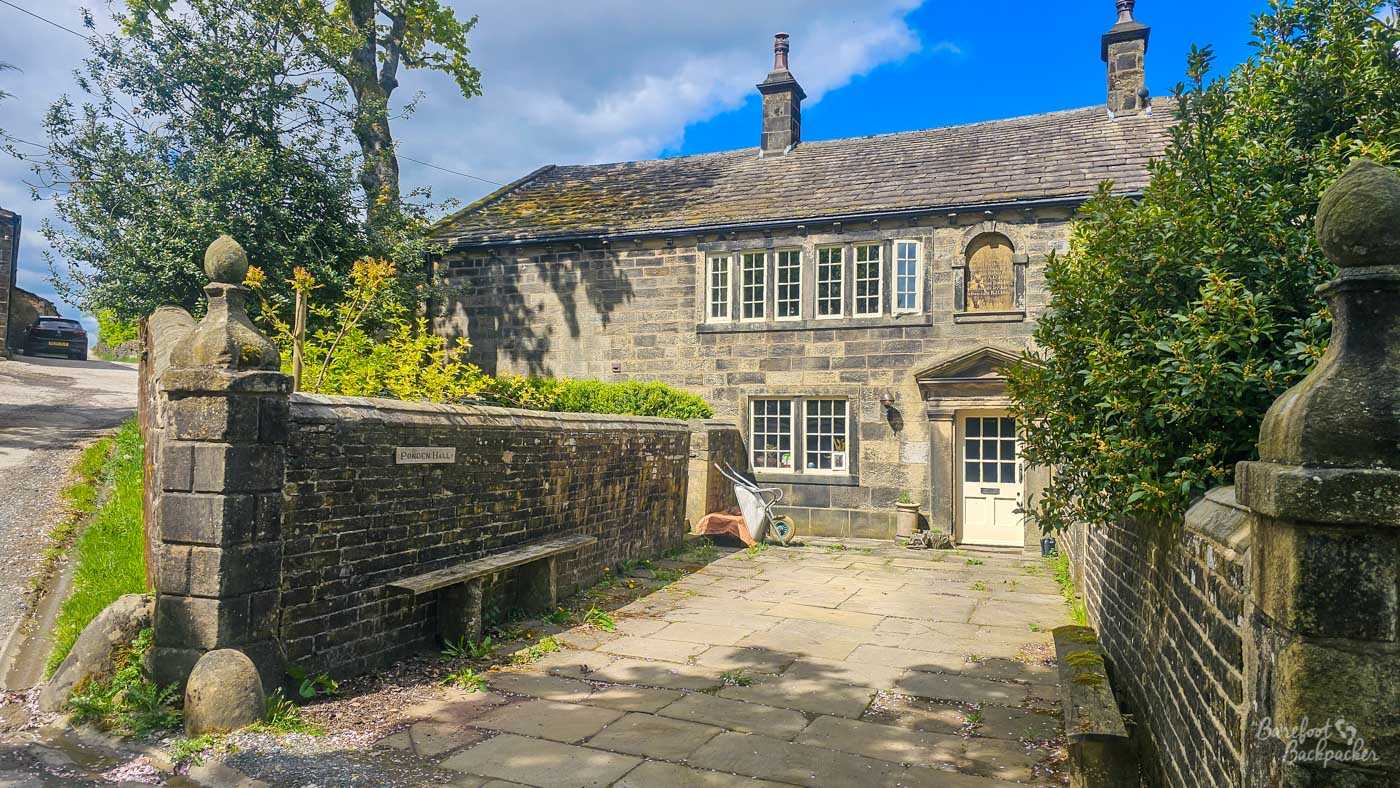 The stone façade of historic Ponden Hall is viewed across its small paved courtyard, with mullioned windows, a central doorway and high boundary walls framing the entrance.