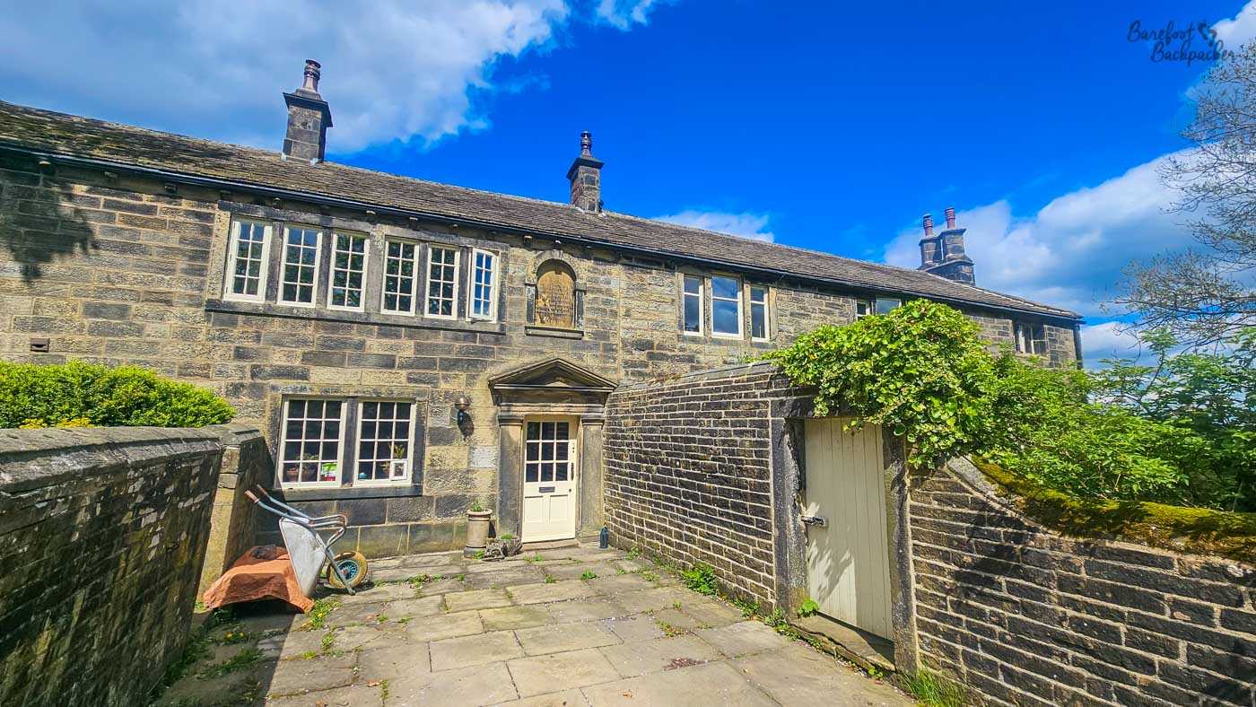 A wide view of Ponden Hall shows the historic stone house behind its enclosed courtyard, bordered by mature trees and traditional stone walls.