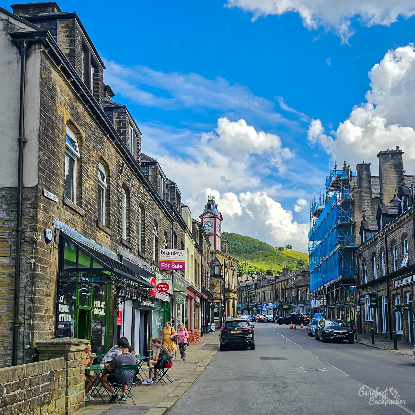 Stone buildings line Marsden's main street beneath a bright blue sky, with the Victorian clock tower standing above shops and cafés against the green hillside beyond.