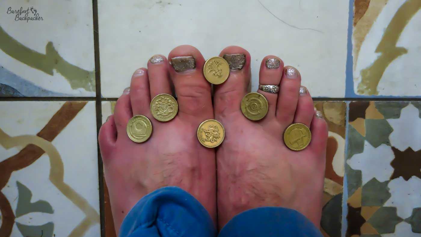 Bare feet with old pound coins balanced on each toe on a patterned tiled floor.