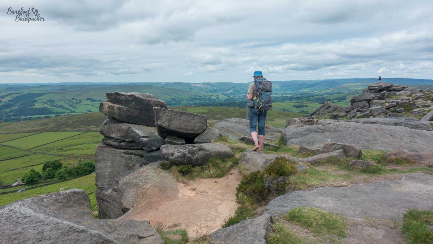 A barefoot hiker stands among the gritstone outcrops of Stanage Edge, looking out across the patchwork fields and rolling hills of the Peak District beneath cloudy skies.