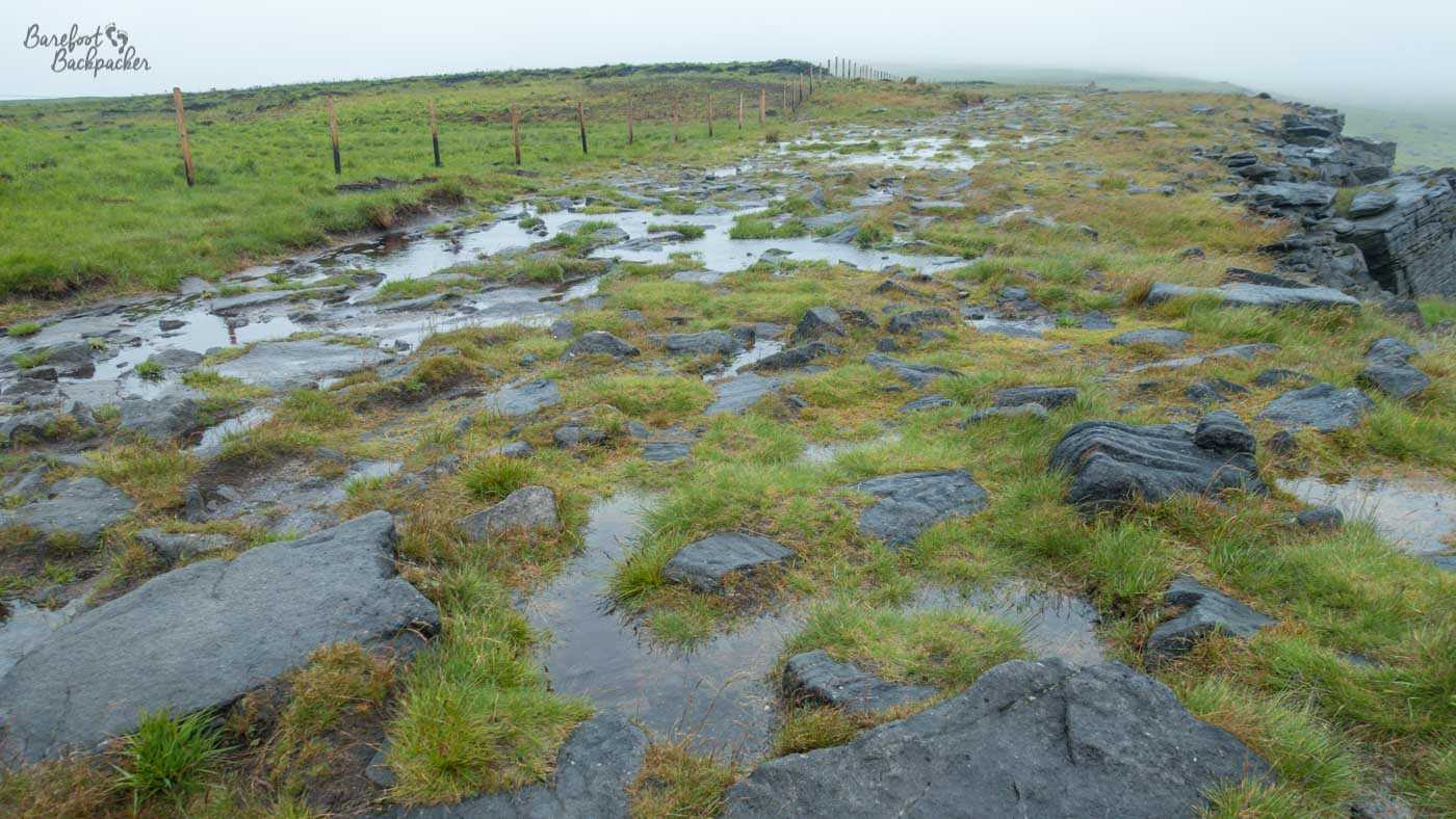 Rainwater pools across a rocky moorland path beside a fence line, with low cloud and mist obscuring the landscape beyond the edge of the escarpment.