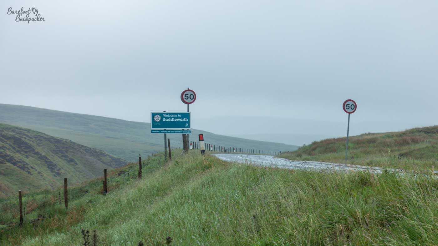 A roadside sign welcoming visitors to Saddleworth stands on a lonely moorland road disappearing into mist, flanked by 50 mph speed limit signs.