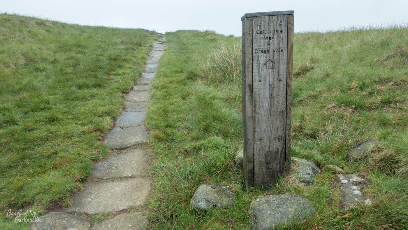 Wooden Calderdale Way waymarker beside a flagstone footpath crossing open moorland, showing the way to Cragg Vale