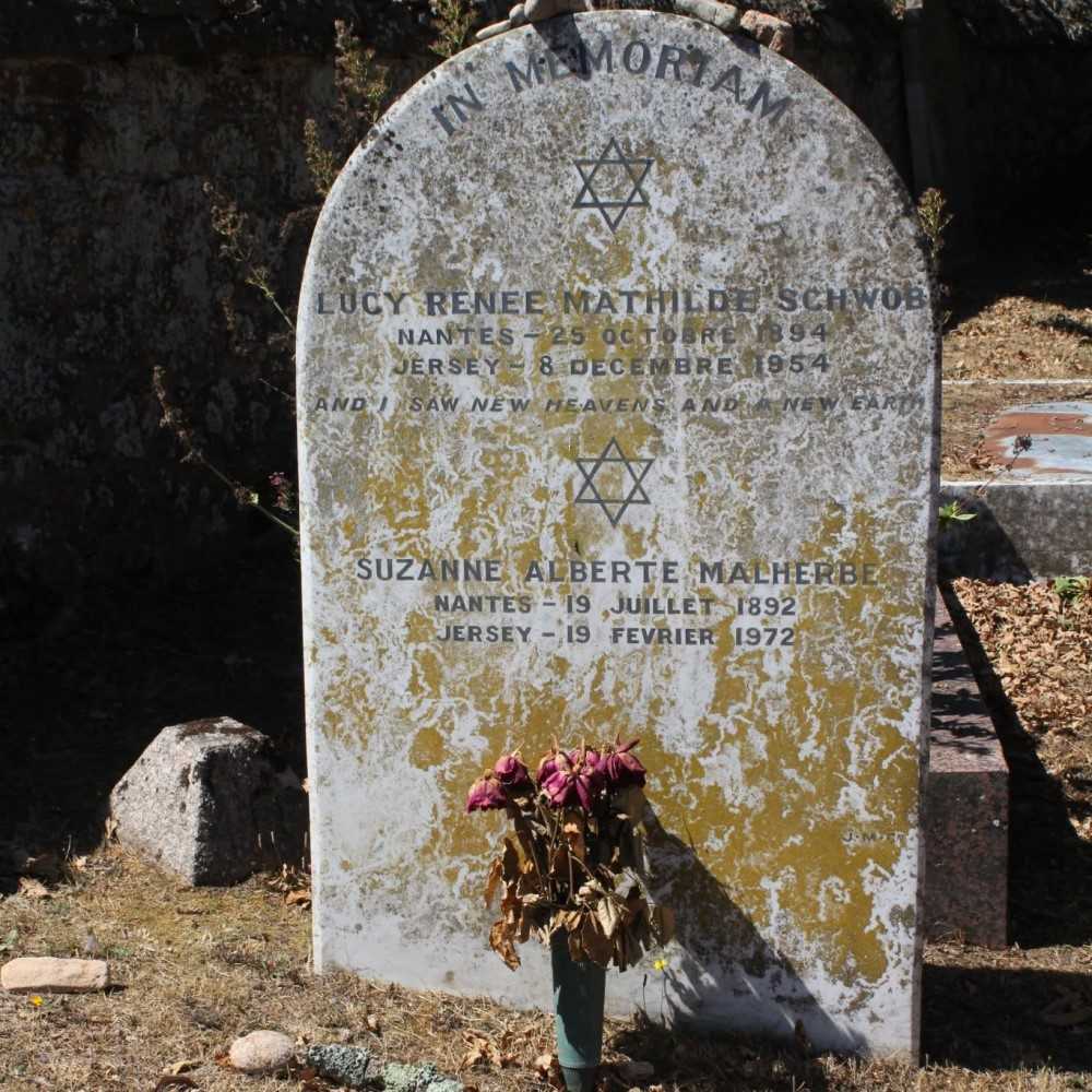 A tombstone reads 'In Memoriam Lucy Renee Mathilde Schwob Nantes - 25 Octobre 1894 Jersey - 8 Decembre 1954 And I Saw New Heavens and a New Earth Suzanne Alberte Malherbe Nantes - 19 Juillet 1892 Jersey - 19 Fevrier 1972.' At the top of the tombstone, there is a Star of David. Below Lucy Schwob's inscription, there is another Star of David. Dried flowers in a vase are at the bottom of the tombstone. The tombstone is weathered and stained yellow and green. The background shows a stone wall and a cemetery setting with dry leaves and grass.