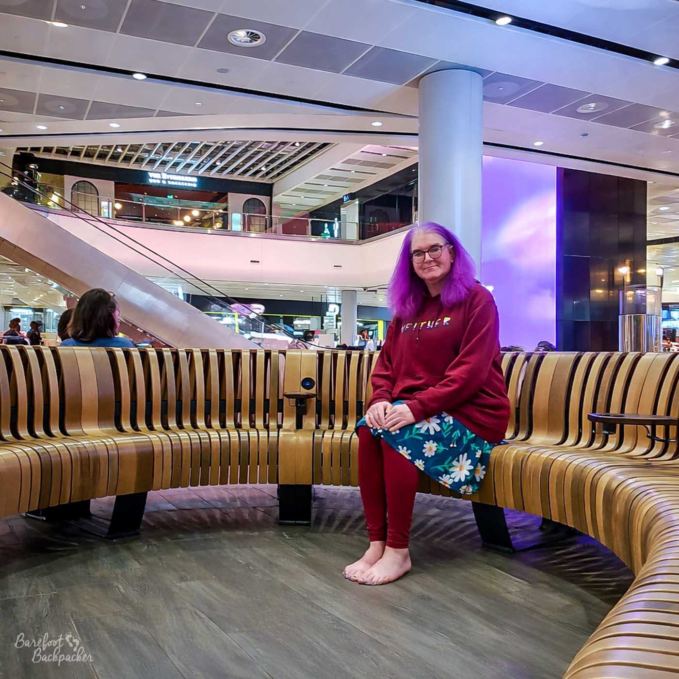 A woman with bright purple hair and glasses smiles at the camera while sitting on a curved wooden bench in a modern indoor space. She is wearing a burgundy hoodie with white lettering that reads